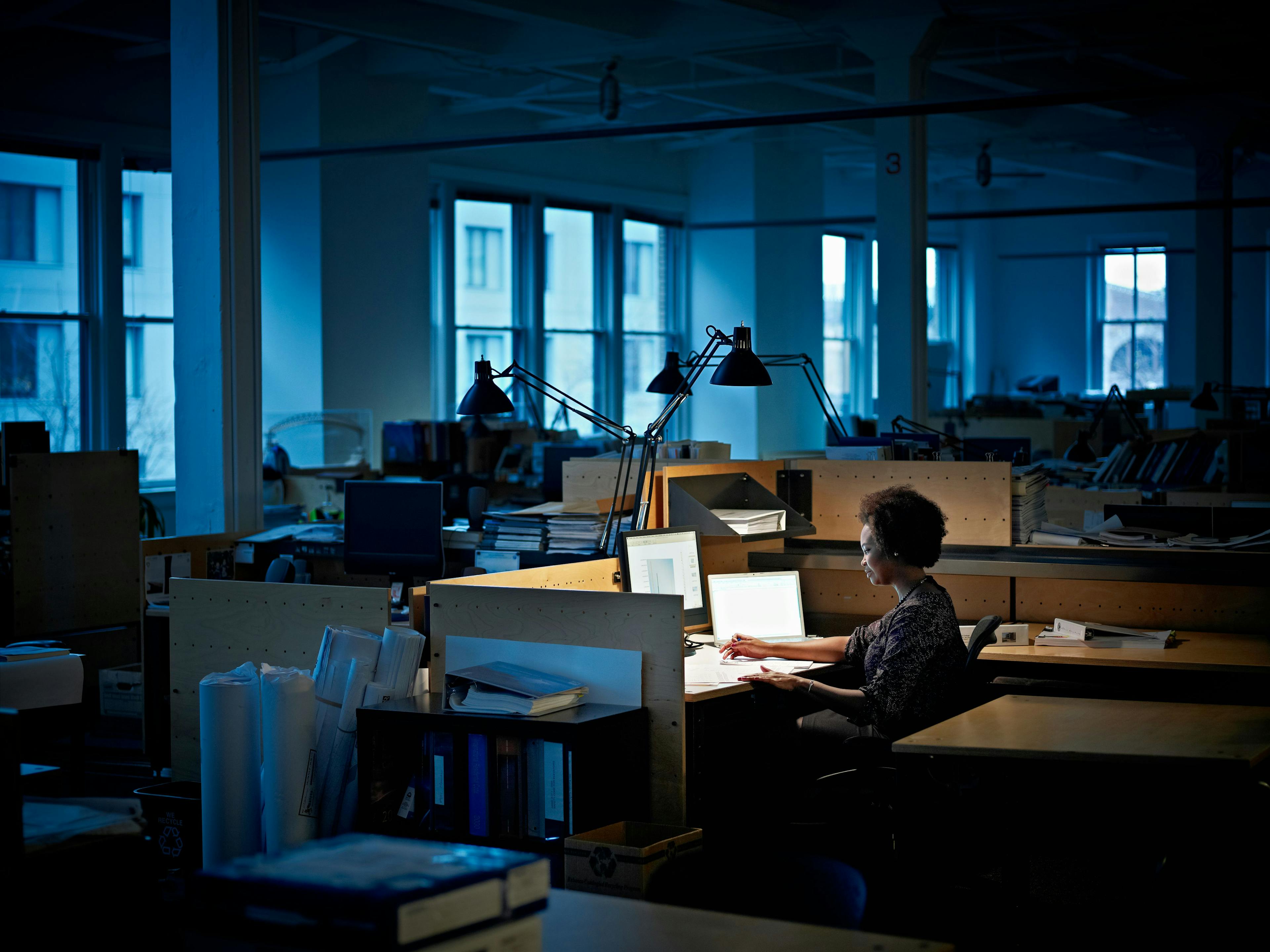 A person sits alone at a desk in a deserted, dimly-lit office.
