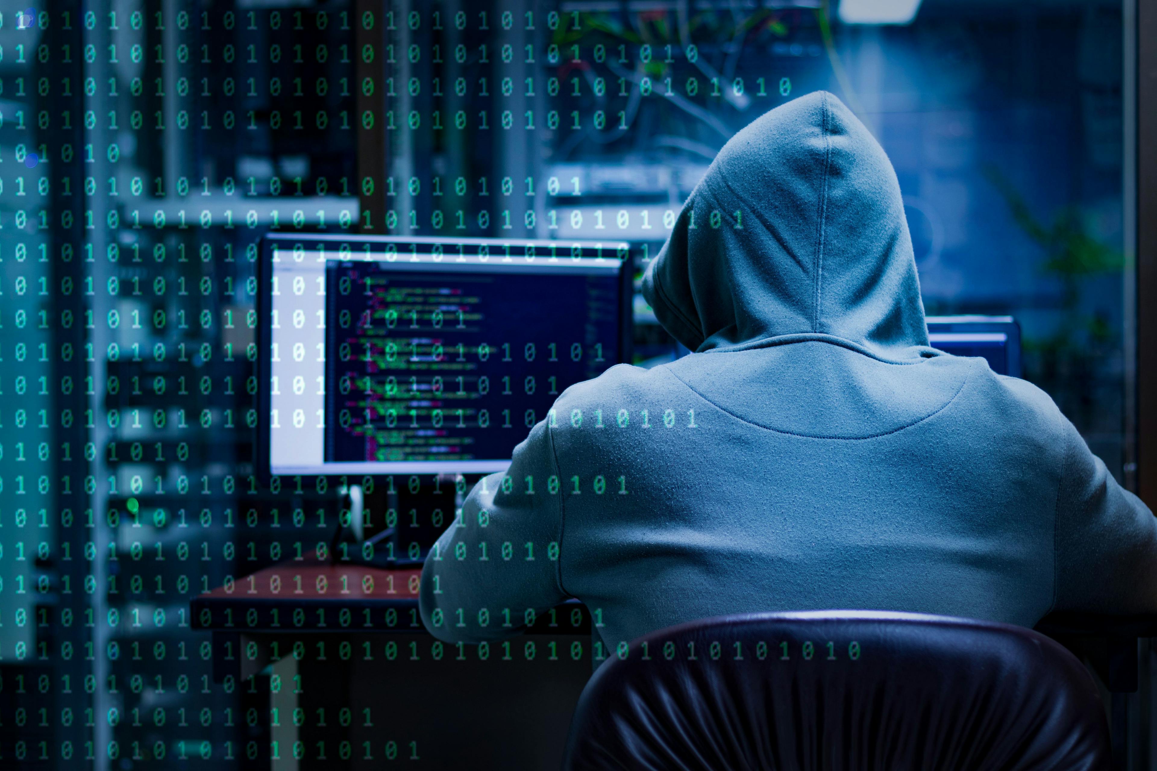 A person in a hoodie types at a computer in a dark room.